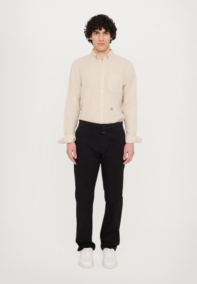 Chemise beige en lin à boutons avec une poche poitrine, associée à un pantalon noir et des baskets blanches, présentée sur un fond neutre.