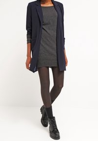 Blazer long bleu marine porté sur une robe grise, associé à des collants noirs et des bottines à lacets. Le blazer comprend des poches ouvertes et une coupe décontractée.