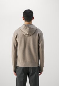 Hellbrauner Kapuzenpullover mit weicher Textur, ausgestattet mit einer Kängurutasche und einem gerippten Saum, kombiniert mit dunkelgrauen Hosen.