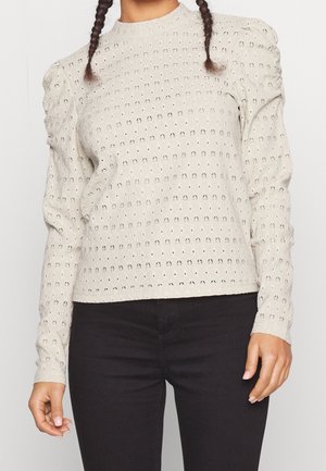 Frau trägt beige texturierte Bluse mit langen Ärmeln und aufgeblasenen Schultern sowie schwarze Hose, steht vor einem schlichten hellen Hintergrund.