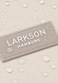 Beige színű textúrázott szövet, vízcseppekkel, egy téglalap alakú szürke címkével, amelyen fehér, dombornyomott betűkkel szerepel a "LARKSON HAMBURG" felirat.
