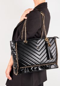Borsa tote nera con motivo a zigzag trapuntato, manici in catena dorata, nappina e bordo arricciato in fondo. Texture liscia e forma strutturata.