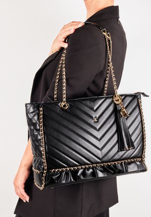 Musta kankainen olkalaukku, jossa on palttinapinta, kullatut ketjuhihnat, tupsu ja röyhelöalasreuna. Sujuva pinta ja jämäkkä muoto.