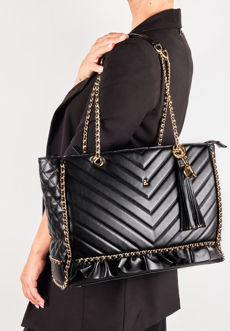 Borsa tote nera con motivo a zigzag trapuntato, manici in catena dorata, nappina e bordo arricciato in fondo. Texture liscia e forma strutturata.