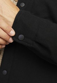 Zwart textiel met een zichtbaar manchet, voorzien van een grote zwarte drukknoop en een binnenlaag van een geruite stof.