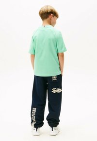 T-shirt a maniche corte verde menta con dettaglio del colletto rosso e bianco, abbinato a pantaloni sportivi blu marino con testo e loghi bianchi "Tommy".