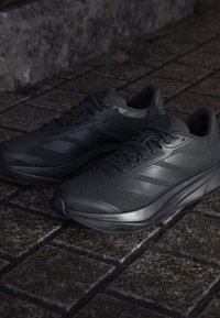 Paire de chaussures de course athlétiques noires sur un trottoir en pierre sombre avec des détails texturés et lacets noués.