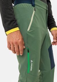 Groene outdoorbroeken met een slank ontwerp, voorzien van ritssluitingzakken in zwart en groen, en een logo op de dij. De stof lijkt soepel en duurzaam.