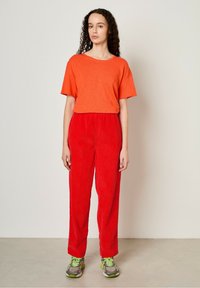 T-shirt orange à manches courtes associé à un pantalon en velours côtelé rouge. Le modèle porte des baskets grises et vertes, debout sur une surface neutre.