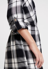 Camicia a quadri bianca e nera con maniche arrotolate, dotata di dettagli con bottoni. Il materiale sembra morbido con una superficie liscia e una vestibilità su misura.