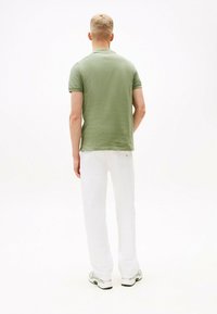 Camisa polo verde de manga corta con un acabado texturizado, combinada con pantalones blancos y zapatillas grises, vista desde la parte trasera sobre un fondo liso.
