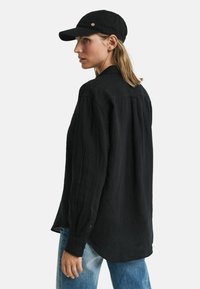 Zwarte linnen overhemd met lange mouwen, iets los model, gecombineerd met een zwarte pet met een ronde gouden accent. Blauwe denim jeans maken de look compleet.