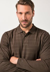 Polo shirt a maniche lunghe marrone scuro con un motivo a strisce sottile, colletto con bottoni e logo accentuato sulla manica. Tessuto morbido e texturizzato.