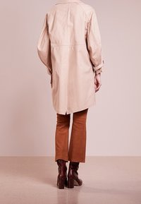 Personne portant un trench-coat beige, un pantalon évasé marron et des bottes à talons marron foncé, debout devant un fond rose clair.
