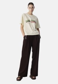 T-shirt di cotone giallo chiaro con stampa grafica; pantaloni larghi neri con borchie decorative alle tasche; sandali con piattaforma marroni.