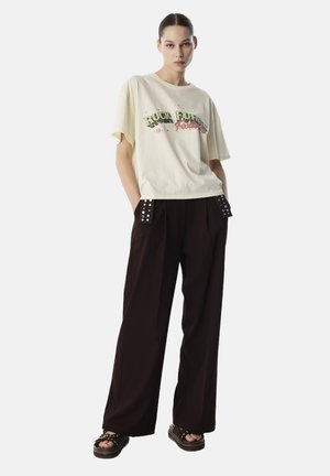 T-shirt en coton jaune clair avec impression graphique ; pantalon large noir avec des clous décoratifs aux poches ; sandales à plateforme marron.