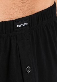 Pantalones cortos negros de algodón ligero con una cinturilla elástica, que cuentan con una pequeña etiqueta circular con logo y un bolsillo lateral. Textura suave.