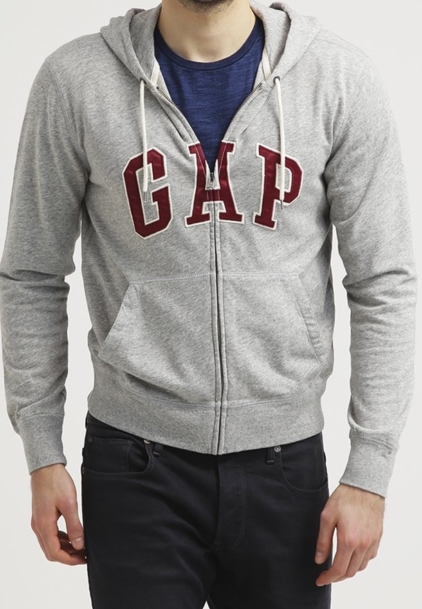 Hombre vestido con una sudadera gris con cremallera que tiene "GAP" en letras rojas sobre una camiseta azul marino, combinado con pantalones negros, de pie frente a un fondo blanco.