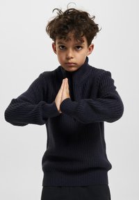 Jūras spēku adīts džemperis ar augstu apkakli un ribotu tekstūru. Tam ir garas piedurknes un pieguļošs viduklis, uzsverot ikdienišķo dizainu.