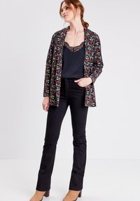 Chaqueta negra con estampado floral sobre una camiseta negra, combinada con jeans acampanados negros y botas de tacón en tono marrón. La chaqueta tiene un corte holgado.