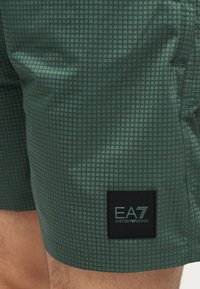 Πράσινα κοντά σορτς με καρό σχέδιο, με μαύρη ετικέτα στο κάτω μέρος του μηρού που γράφει "EA7 Emporio Armani", φορεμένα από ένα άτομο.