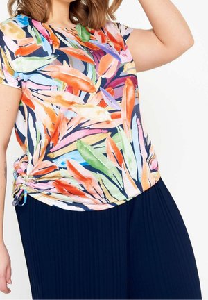 Vrouw draagt een kleurrijke top met korte mouwen en abstracte veerpatronen en marineblauwe geplooide broek tegen een witte achtergrond.