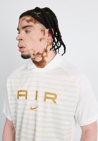 White polo shirt with horizontal golden stripes featuring "AIR" in large font. Soft fabric with a classic collar and short sleeves.