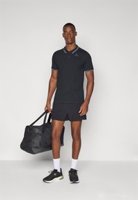 Svart piké med hvit kant, kombinert med svarte shorts. Svart duffelbag båret i en hånd, og svarte treningssko med hvite sokker.