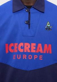 Modrá polo košeľa s námorníckym golierom, s veľkým červeným logom "ICECREAM EUROPE" a vyšívanou postavou farebnej zmrzliny.