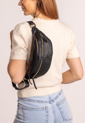 Bolso de cuero negro tipo bandolera con dos cremalleras frontales, correa ajustable, forma curvada y textura suave, llevado al hombro sobre un fondo claro.