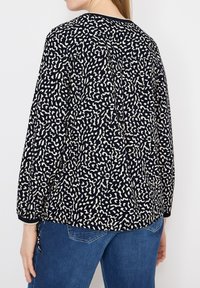 Femme portant un chemisier à manches longues à imprimé abstrait noir et blanc avec un col rond, associé à un jean en denim bleu, vue de dos.