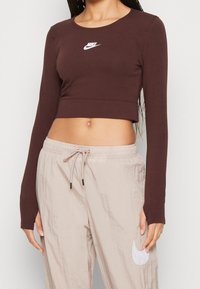 Brun långärmad crop top tillverkad av stretchigt material, med en vit Nike-logga på bröstet, i kombination med ljusbeige byxor med elastisk midja.