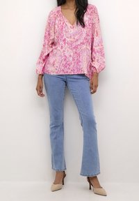 Blusa paisley rosa con scollo a V e maniche a palloncino, abbinata a jeans chiari a zampa d'elefante e scarpe con tacco nude. Texture liscia.