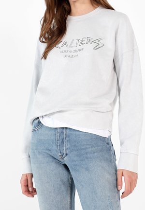 Frau trägt hellgrauen Pullover mit dem Aufdruck "Scalpers Always On Fire" und Sternen, kombiniert mit hellblauen Jeans.