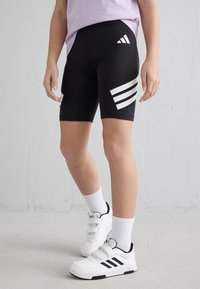 Mustat urheilushortsit, joissa on valkoiset kolmiraidalliset yksityiskohdat sivuissa, valmistettu sileästä ja joustavasta materiaalista. Yhdistetty valkoisten lenkkareiden ja sukkien kanssa.