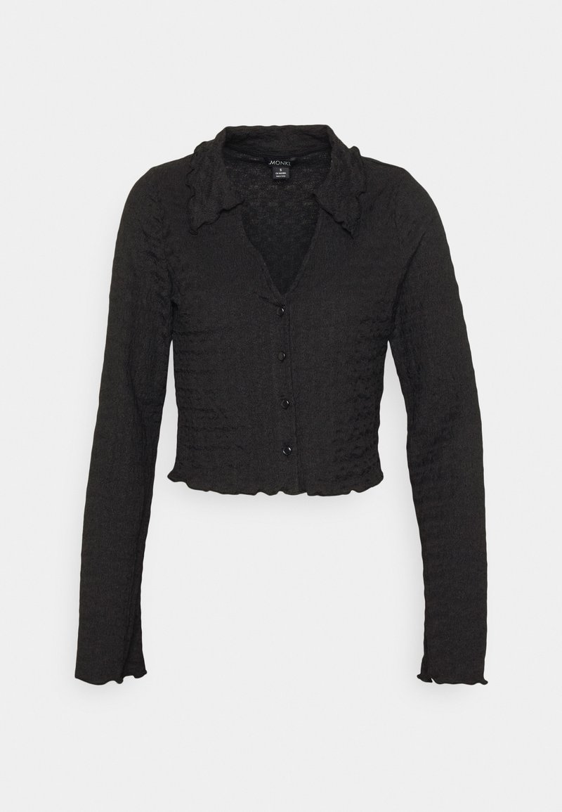 Cárdigan corto negro de tejido texturizado, con mangas largas y un cuello con volantes. Cinco botones en la parte delantera añaden detalle.