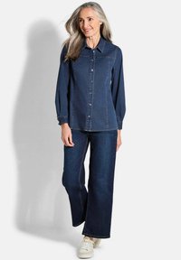 Dunkelblaus Denimhemd mit Knopfleiste, spitzem Kragen und langen Ärmeln; kombiniert mit weit geschnittenen dunklen Jeans und weißen Sneakern.