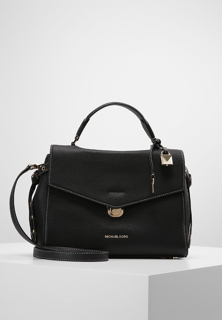 Sac à main Michael Kors en cuir texturé noir avec poignée supérieure, fermoir doré, breloque en forme de cadenas et bandoulière amovible sur un piédestal blanc.