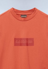Kurzärmeliges T-Shirt aus leuchtend orangefarbenem Baumwollstoff, mit weicher Textur und einem vorderen Grafiklabel in einer dunkleren Farbe mit dem Markennamen.