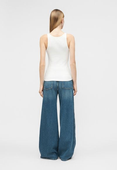 Γυναίκα με μακριά ξανθά μαλλιά, που φοράει λευκό αμάνικο τοπ και φαρδιά μπλε τζιν παντελόνια, στέκεται με την πλάτη σε έναν απλό φόντο.