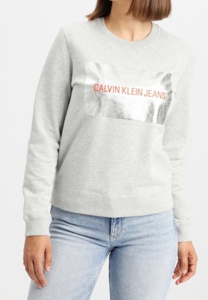 Sweat-shirt gris en mélange de coton, présentant un graphisme métallique argenté avec "CALVIN KLEIN JEANS" en lettres rouges sur le devant.