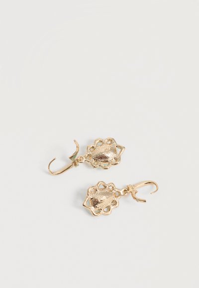 Κρεμαστά σκουλαρίκια σε χρυσό τόνο με υφή, οβάλ κρεμαστά στοιχεία με κυματιστές άκρες και αγκίστρους, σε λευκό φόντο.