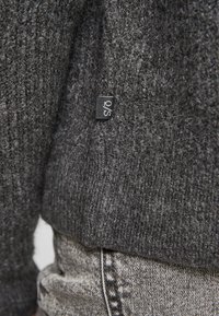 Donkergrijze gebreide trui met zichtbare textuur, voorzien van een klein zwart label met witte tekst. Gecombineerd met lichtgrijze jeans.