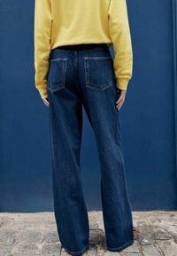 Persona, kas valkā platām kājām tumši zilas džinsas un dzeltenu džemperi, stāv uz bruģakmens pret zilu sienu.