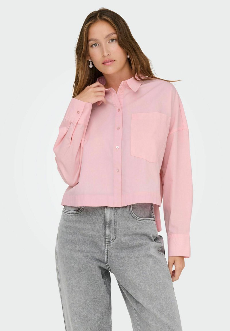 Camicia rosa chiaro a maniche lunghe, cropped, con un taschino sul petto e una morbida texture del tessuto, abbinata a jeans grigi a vita alta.