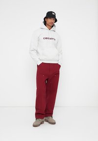 Svetlo šedá mikina s kapucňou a logom "Carhartt" v tmavej marhuľovej farbe, skombinovaná s voľnými marhuľovými nohavicami a šedými topánkami. Čierny klobúk na vedro dotvára celý vzhľad.