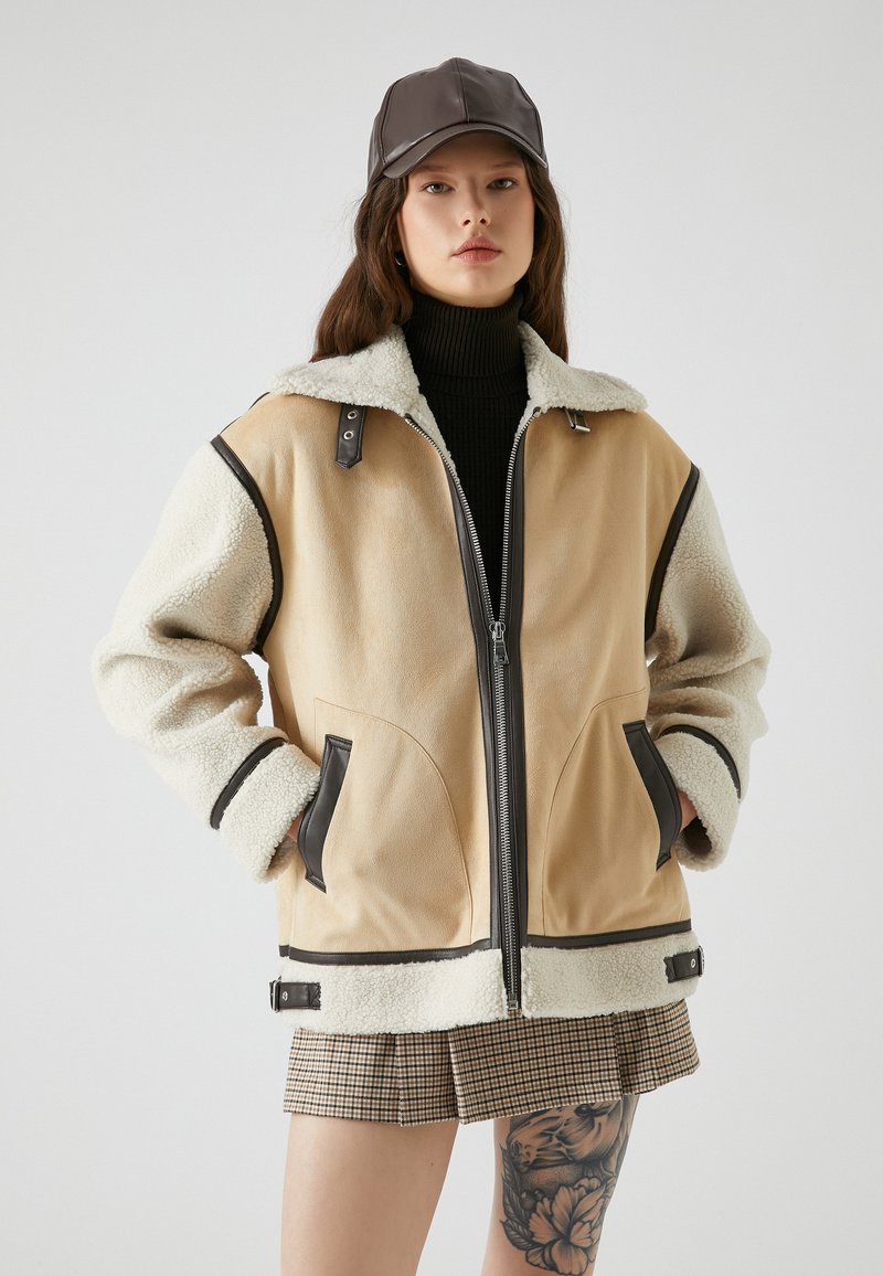 Tann- und cremefarbener Blazer mit kuscheligem Schaffell, schwarzen Lederakzenten, durchgehendem Reißverschluss und seitlichen Taschen. Tragen über einem schwarzen Rollkragenpullover.