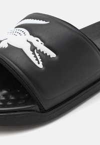 Sandalia de dedo negra de goma con un patrón texturizado, una correa ancha con un logo de cocodrilo blanco y una suela plana para mayor comodidad.