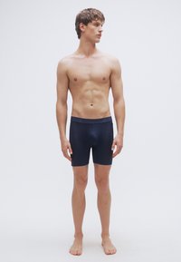 Σκούρα ναυτικού χρώματος στενά μαγιό με φαρδύ λάστιχο στη μέση, λεία υφή και χωρίς ορατά σχέδια. Το μοντέλο στέκεται ξυπόλυτο μπροστά σε ανοιχτό φόντο.