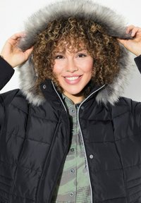 Chaqueta acolchada negra con capucha forrada de piel sintética gris. La chaqueta presenta un diseño acolchado y un cierre frontal con cremallera, llevada sobre una camisa verde con estampado.